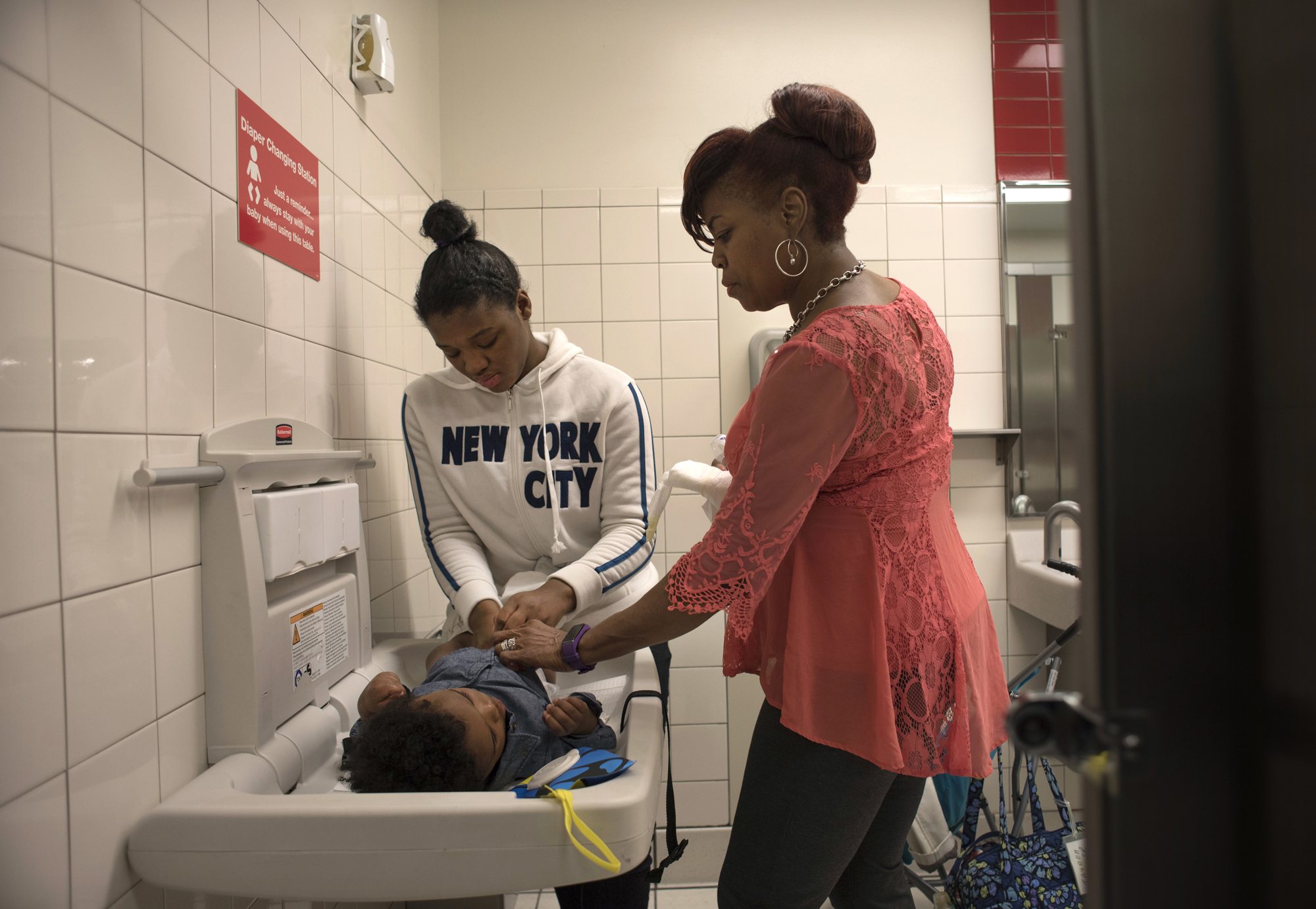 Adrienne Bradshaw, (right) helps Tyneisha Wilder, age 18, change Tayden's diaper in the restroom at Target in East Liberty during one of their twice-weekly supervised visits. (Photo by Martha Rial/PublicSource)